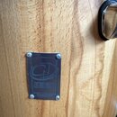 Detail dřevěného povrchu s viditnou masivní strukturou. Na dřevo je čtyřmi šrouby připevněn kovový obdélníkový štítek s vyrytým logem a nápisem CUSTO…