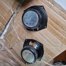 Dvě černé reproduktorové jednotky s kovovými magnety, umístěné na vlnitém kartonu. Na stříbrném povrchu magnetů jsou viditelné bílé nálepky s červený…