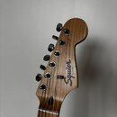 Detail hlavy elektrické kytary z hnědého dřeva s viditnou masivním kresbou. Na hlavě je bílý nápis Squier STRATOCASTER a menší text BY FENDER. Jsou z…