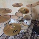 Bílá bicí souprava s několika činely na kovových stojaních. Na bubnech a činelech jsou viditelné nápisy BICS, Meinl a px t. Povrch blan bubnů vykazuj…