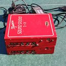 Dvě červené kovové krabičky s nápisy „Mozart SOUND STUDIO 1-X“. Na horní straně je bílý rámeček a v pravém dolním rohu malý štítek s nečitelným texte…