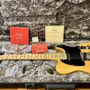 Elektrická kytara typu Telecaster v přirozené dřevěné barvě s černým pickguardem leží v otevřeném tvrdém kufru s šedým plyšovým vnitřkem. Na hlavě ky…