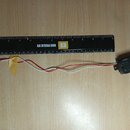 Černý humbucker s dvěma řadami pólů a připojenými červeným a bílým vodičem. Na boku snímače je viditelný nápis EMG. Vedle nástroje leží černá pravítk…