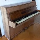 Hnědé dřevěné piano s klaviaturou. Nástroj vykazuje známky opotřebení, zejména v podobě odřenin a vybledlých míst na levém bočním panelu. Na klaviatu…