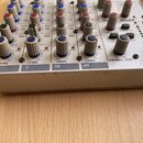 Detailní pohled na horní panel bílého mixážního pultu s řadou ovládacích prvků. Jsou vidět modré, červené a šedé otočné knoflíky s nápisy jako LEVEL,…