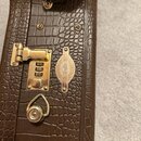 Detail hnědého kytarového kufru s povrchem imitujícím krokodýlí kůži. Na kufru je zlatý zámek a oválný kovový štítek s nápisy Taylor, GUITARS Co. a C…