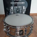 Stříbrný snare drum s kovovým korpusem a bílým blátcem. Na korpusu je umístěn černý čtvercový štítek s logem dvou kuliček a nápisy KOMPRESSOR a SONOR…