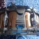 Detailní pohled na část snare bubnu v měděné barvě s chromovaným obručem a lugy. Na držáku mechanismu je vyryt nápis GRETSCH. Povrch nástroje je glos…