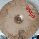 Kruhový činel z mosazi s viditelným opotřebením povrchu a tmavšími skvrnami. V pravé horní části je červený nápis PAISTE a pod ním menší text MADE IN…