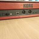 Detail zadní strany hudebního nástroje s dřevěným povrchem v červenohnědé barvě. Na šasi je nápis KORG a panel s konektory, včetně USB portu, dvou MI…