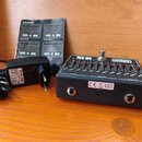 Černý kytarový pedál s nápisy MXR a EQ. Na horní straně jsou posuvníky, spínač a nápisy INPUT, POWER a OUTPUT. Vedle pedálu je černý síťový adaptér a…