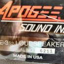 Detailní záběr na štítkování černého zařízení. Je zde vidět červeno-černé logo APOGEE a nápis SOUND INC. Nižší bílý text uvádí model AE-3s2 LOUDSPEAK…