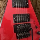 Červená elektrická kytara s dvěma černými humbuckery. Na kobylce je viditelný nápis Floyd Rose a je k ní připojena páka. Povrch nástroje je glossy, s…
