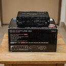 Černý USB audio interface Roland DUO-CAPTURE EX položený na své originální krabici. Na zadním panelu zařízení jsou vidět konektory DC IN, OUT, MIDI I…