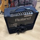 Černý kombozesilovač s tmavě modrým textilním potahem a plastovou rukojetí. Na předním panelu je nápis Roland KC-150 a několik ovládacích knoflíků. N…