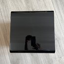 Černý, lesklý předmět čtvercového tvaru s hladkým povrchem. Na horní ploše není žádný viditelný nápis ani logo. V reflexi povrchu je vidět osoba s mo…