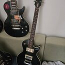 Černá elektrická kytara s tvarem singlecut, bílým lemováním a hnedým hmatníkem. Na hlavě je bílý nápis evo. Kytara má dva humbuckery, čtyři ovládací…