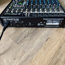 Černý mixážní pult s modrými posuvníky a bílými popisky. Na zadním panelu je nápis ProFX12D 12-CHANNEL PROFESSIONAL EFFECTS MIXER a označení A01 CE.…