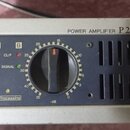 Detailní pohled na přední panel šedého výkonového zesilovače s nápisem POWER AMPLIFIER P2500S. Zařízení má dva velké černé oválné ovladače s červeným…