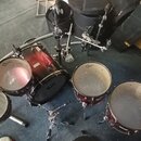 Sada červených bubnů s kovovým kováním. Sestava obsahuje basový buben, dva tom-tomy a jeden floor tom, všechny s bílými membránami. Na membránách je…