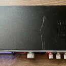 Černý rackový zesilovač nebo procesor s hliníkovým stříbrným rámem. Na předním panelu jsou umístěny ovládací knoflíky, červené kontrolky a modře pods…