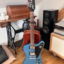 Modrá elektrická kytara s černým hmatníkem a hlavou, umístěná na černém stojanu. Na hlavě je bílý nápis Gibson. Tělo kytary má modrý lak, dva snímače…