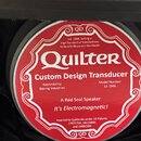 Detailní záběr na červenou kulatou nálepku s bílým textem a ornamenty. Na nálepce je velký nápis Quilter a text Custom Design Transducer, Model Numbe…