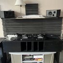 Na fotografii je set DJ vybavení umístěné na černých akustických panelech a bílém stolku. V horní části jsou dva černé studiové monitory, vprostřed č…