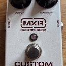 Stříbrný efekt pedál s nápisy MXR CUSTOM SHOP a CUSTOM COMP. Na horní straně jsou dva černé oválné ovládací knoflíky s popisky OUTPUT a SENSITIVITY.…