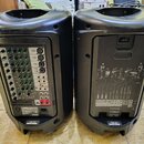 Dva černé plastové aktivní reproduktory z pohledu zezadu. Levý reproduktor má viditelný panel s konektory, ovládacími prvky a nápisy YAMAHA, STAGEPAS…