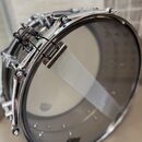 Detailní pohled na chromovaný obruč a napínací mechanismus bubnu s průhlednou koží. Na koži je připevněn bílý textilní tlumič (snare dampener). Na po…