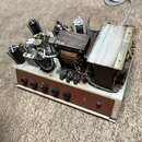 Fotografie zachycuje otevřený elektronický přístroj, pravděpodobně zesilovač, s viditelným vnitřním zapojením. Uvnitř jsou umístěny elektronky, dva v…
