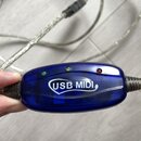 Modrý průhledný plastový adaptér s nápisem USB MIDI. Na horní straně jsou tři kontrolky v barvách zelené, žluté a červené. Adaptér je připojen k stří…