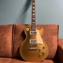 Zlatá elektrická kytara s tmeleým hnedým hmatníkem a bílými bindingy. Na hlavě je viditelné logo Gibson. Kytara má dva humbuckery, čtyři ovládací kno…