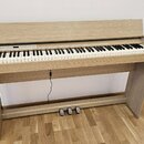 Digitální piano v dřevěném provedení světlé barvy s přívodním kabelem a třemi pedály. Na pravém boku korpusu je nápis "estland". Nástroj má bílé a če…