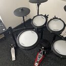 Elektronická bicí souprava černé barvy s bílými membránami. Na bubnech jsou viditelné nápisy Roland a V-Drums. Souprava obsahuje dva činely, čtyři pa…