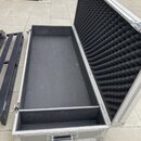 Otevřený hliníkový transportní kufr s černým vnitřním povrchem. Víko je vyplněno černou akustickou pěnou s vlnovitým profilem. Kufr má stříbrné kovov…