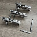 Tři kovové stříbrné svorky s nápisy FastClamp TAMA. K nim je přiložen jeden L-klíč z kovového materiálu. Všechny předměty leží na dřevěném povrchu.