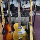 Tři elektrické kytary v držáku před kytarovým kombem značky Marshall. Uprostřed je zlatá polodupatá kytara s nápisem GRETSCH na hlavě a nápisem G6120…