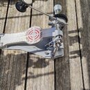 Fotografie zachycuje kovový mechanismus bubnového pedálu v stříbrné barvě, umístěný na dřevěném podkladu. Na bočním členu je červené kulaté logo s ná…