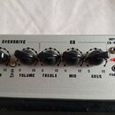 Detail ovládacího panelu zesilovače v stříbrné barvě. Panel obsahuje šest černých otočných ovladačů s nápisy GAIN, VOLUME, TREBLE, MID a BASS, vstup…