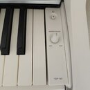 Detailní pohled na ovládací panel bílého digitálního pianina. Na panelu je umístěno tlačítko napájení, otočný ovladač s nápisy MASTER VOLUME, MIN a M…