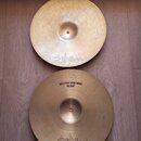 Dvě mosazné činely typu hi-hat položené na dřevěné podlaze. Oba mají v dolní části nápis Zildjian. Spodní činel má navíc v horní části nápis BOTTOM N…
