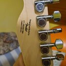 Zadní strana světlé dřevěné hlavy basové kytary s šesti chromovanými ladičkami. Na dřevě je černý nápis „Designed and Backed by Fender“, pod ním „s/n…