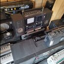 Fotografie zobrazuje několik kusů starší audio techniky v černém provedení, včetně boomboxů a hi-fi systémů. Na horním zařízení je viditelný nápis PC…