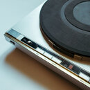Stříbrný gramofon s černým talířem. Na ovládacím panelu jsou vidět nápisy REPEAT, SIZE, SPEED, TRACK NUMBER, 50Hz, 60Hz, 33 RPM, TRACK a SKIP. V levé…