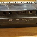 Detail zadní části černého klavírního nástroje s řadou konektorů. Jsou zde nápisy SUSTAIN, PHONES/OUTPUT, AUX IN, TO DEVICE, USB s ikonou USB a TO HO…