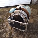 Černý transportní flightcase s hliníkovými hranami a dvěma madly, který stojí na kolečkách. Uvnitř jsou uloženy dvě velké cívky s šedým kabelem, kter…