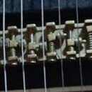 Detailní záběr na mosazný kobylník elektrické kytary s šesti sedly. Kovový povrch vykazuje známky oxidace a opotřebení. Na černém povrchu kytary je v…