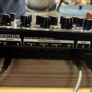 Zadní panel černého analogového syntetizátoru s nápisy MINITAUR a analog bass synthesizer. Jsou zde vidět vstupy a výstupy s popisky AUDIO (OUT, IN),…